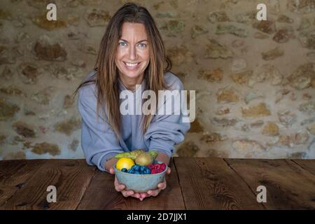 Sorridente nutrizionista femminile con vari frutti in ciotola seduta a. tavolo contro muro di pietra Foto Stock