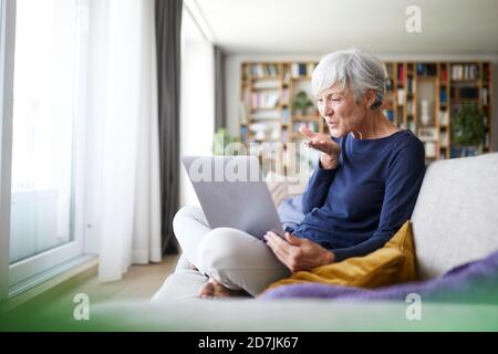 Donna anziana che dà bacio volante in videochiamata mentre si è seduti a casa Foto Stock