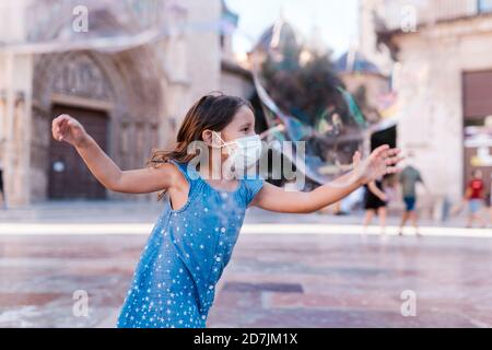 Ragazza spensierata indossando maschera che corre su strada in città giocando con bolla di sapone Foto Stock