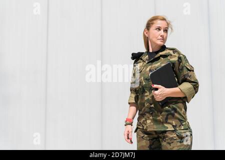 Soldato dell'esercito biondo maturo che tiene un tablet digitale mentre si guarda via alla base militare in giornata di sole Foto Stock