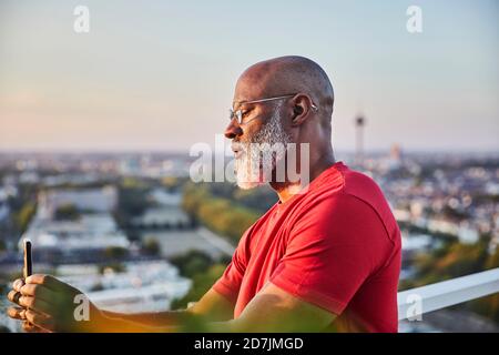 Uomo calvo con barba bianca che usa lo smartphone per fotografare tramonto sulla città dalla terrazza Foto Stock