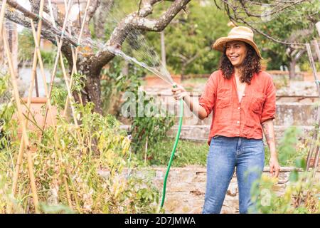 Sorridente bella donna matura annaffiando piante da tubo in giardino Foto Stock