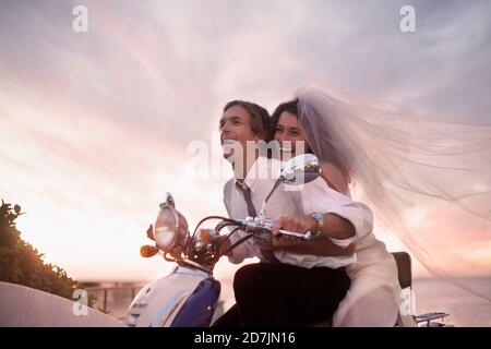 Allegra coppia newlywed in abito da sposa guidando scooter durante tramonto Foto Stock