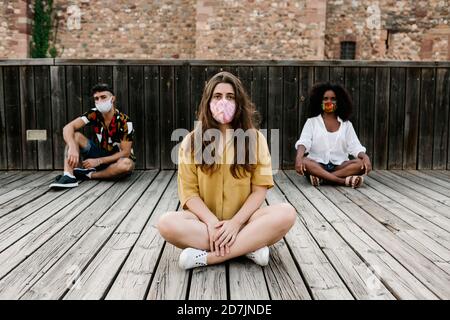 Gli amici indossano una maschera protettiva mentre si siedono su un pavimento in legno durante la covid-19 Foto Stock