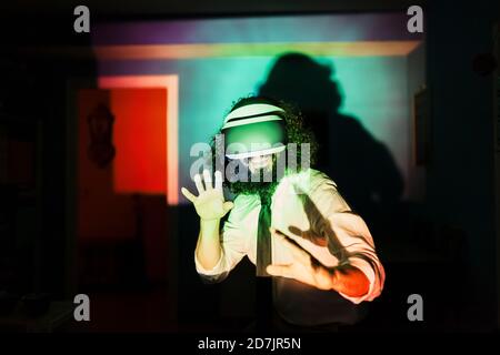 Uomo sorridente che indossa occhiali per realtà virtuale in piedi multicolore riflessione della luce a casa durante la pandemia Foto Stock