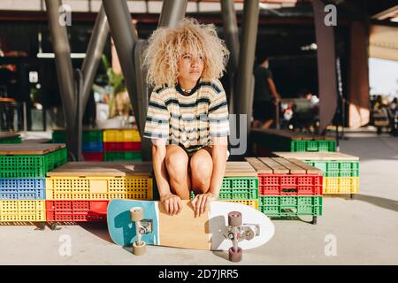 Donna bionda afro che tiene lo skateboard e guarda via mentre si siede sul sedile in cassa durante la giornata di sole Foto Stock