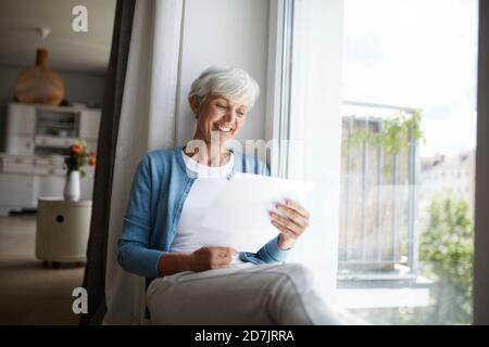 Donna anziana che legge la lettera felicemente mentre si siede sulla finestra a. casa Foto Stock