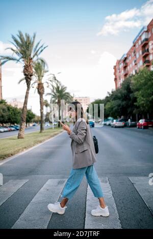 Giovane donna che usa lo smartphone mentre cammina sulla traversata di zebra Foto Stock