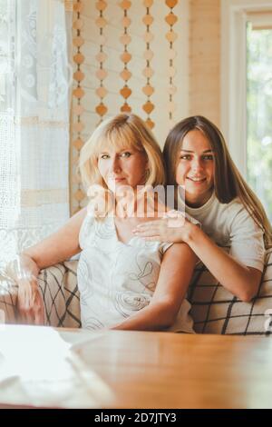 Amorevole adulto 20s figlia abbraccio anziana madre da dietro mentre mamma seduta sul divano persone che posano guardando la fotocamera sorridente si sente felice, concetto di mu Foto Stock