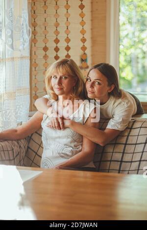 Amorevole adulto 20s figlia abbraccio anziana madre da dietro mentre mamma seduta sul divano persone che posano guardando la fotocamera sorridente si sente felice, concetto di mu Foto Stock