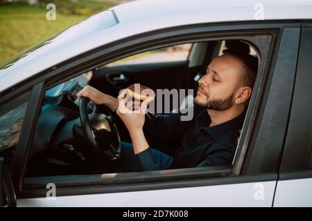 Un bel giovane uomo d'affari caucasico seduto in una macchina, andando a lavorare e mangiare una deliziosa ciambella con crema di cioccolato. Un pasto dolce in auto Foto Stock