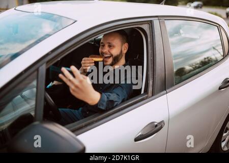Un bell'uomo caucasico sorridente che prende un selfie e mangia una ciambella con cioccolato mentre è bloccato nel traffico. Un dolce pasto di un uomo d'affari Foto Stock