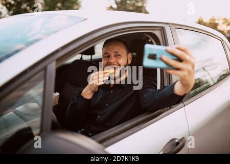Un uomo caucasico sorridente che prende un selfie mentre mangia una ciambella con cioccolato in macchina. Un pasto dolce in auto Foto Stock