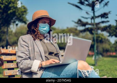 Donna lavora seduto in un parco e si protegge con una maschera medica. Ritratto di una donna alla moda che ama gli spazi aperti e scrive sul suo blog Foto Stock