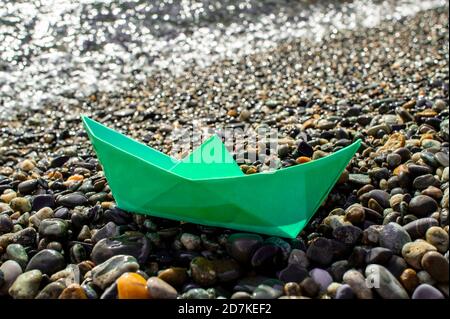 Green paper boat primo piano di una spiaggia di ciottoli sullo sfondo del mare. Origami. Design poster Foto Stock