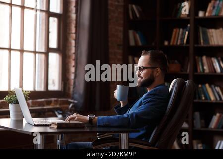 Caffè fresco al lavoro. Giovane sicuro in abbigliamento casual elegante che lavora su un computer portatile e beve una bevanda calda mentre si siede al lavoro Foto Stock