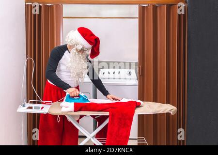 Babbo Natale si prepara per Natale, stirando i suoi vestiti nella lavanderia. Stirare la giacca rossa sul ferro da stiro Foto Stock