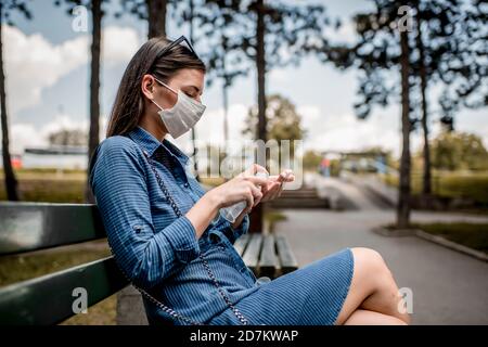 Giovane donna che indossa una maschera medica per il viso e che usa una mano con alcol igienizzatore per pulire le mani Foto Stock