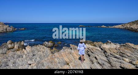 Panorama di donna vista posteriore in un abito blu si erge su una riva rocciosa e si affaccia sul mare in una giornata limpida. Spazio di copia Foto Stock