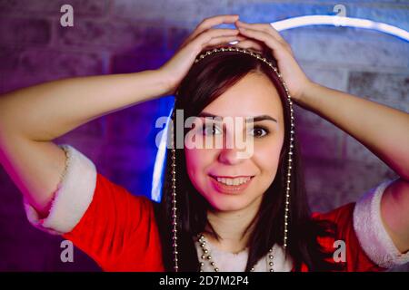 Ritratto di giovane donna in abito di Babbo Natale con perline contro parete illuminata. Primo piano di bella femmina in costume di Natale tenuta perline. Concetto Foto Stock