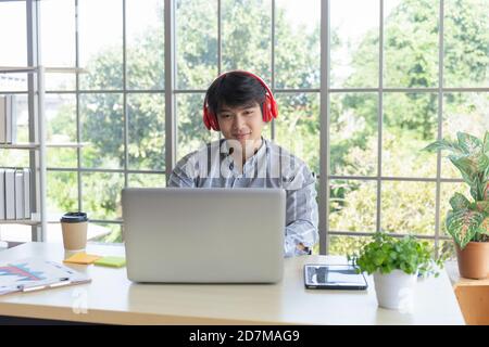 Un giovane uomo d'affari asiatico ascolta la musica su una scrivania su un balcone con finestra di vetro. Foto Stock