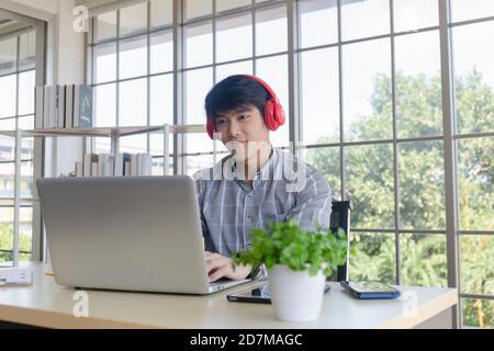 Un giovane uomo d'affari asiatico ascolta la musica su una scrivania su un balcone con finestra di vetro. Foto Stock