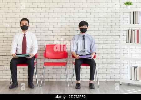 Due uomini asiatici si siedono in sedie di plastica rossa distanziate con Matt per coprire la bocca per evitare COVID-19 in attesa di un colloquio di lavoro. Foto Stock