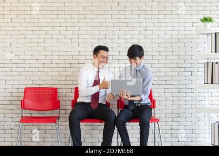Due uomini asiatici siedono su sedie di plastica rossa in attesa di un colloquio di lavoro. Foto Stock