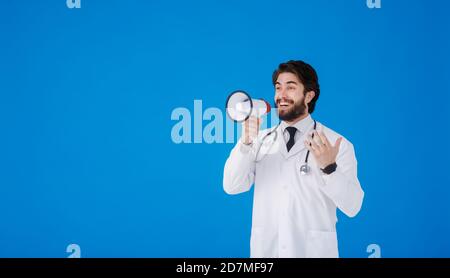 un giovane medico bearded su uno sfondo blu con un il megafono nelle sue mani grida Foto Stock