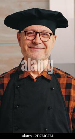 Vecchio uomo caucasico che indossa grembiule in cucina casalinga sorridendo alla macchina fotografica. Panettiere anziano in pensione con bonete preparazione di ingredienti di pasticceria su tavola di legno pronto a cucinare pane, torte e pasta gustosi fatti in casa. Foto Stock