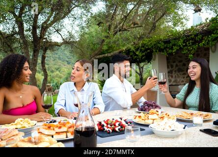 Multi razza felice gruppo di amici giovani tostatura in giardino Al tramonto con bicchieri di vino rosso - millennials bonding e. parlare al tavolo carico in paese Foto Stock