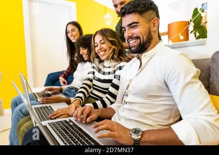 Gruppo di giovani amici multietnici a casa seduti Il divano con tre computer portatili in alluminio - vista laterale di ha allineato i millennial lavorando e divertendosi Foto Stock