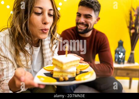 Giovane bella donna bionda soffia fuori la candela di lei torta di compleanno con il suo ragazzo sorridente seduto sul divano - Celebrazione di coppia a casa - Foto Stock