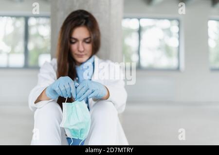 Triste e disperata giovane dottoressa che tiene una maschera protettiva seduta sul pavimento in ospedale dopo il turno. Concetto di coronavirus covid-19. Foto Stock