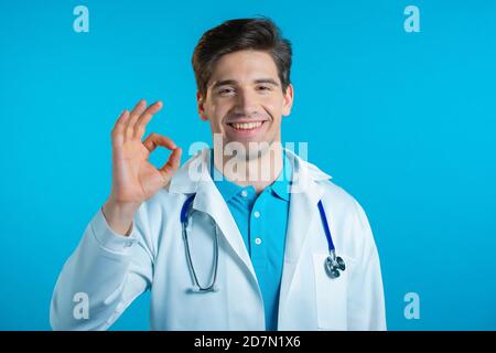 Uomo in camice medico bianco che mostra OK segno su sfondo blu. Il medico positivo sorride alla macchina fotografica. Vincitore. Operazione riuscita. Linguaggio del corpo. Foto Stock