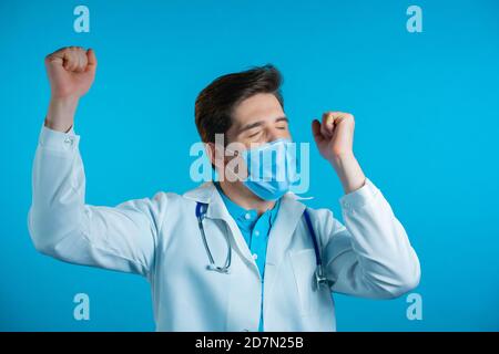 Sì gesto vincitore. Stupito medico in bianco cappotto professionale scioccato. Bell'uomo sorpreso a macchina fotografica su sfondo blu Foto Stock