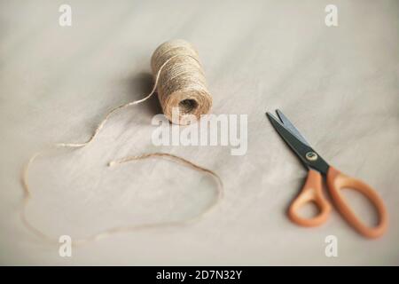 Sul tessuto beige si trova una bobina di corda di canapa e moderne forbici. Processo di fabbricazione. Hobby. Foto Stock