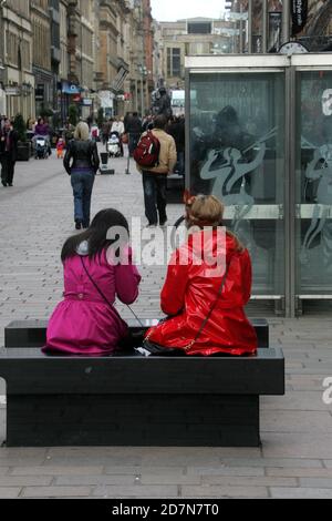 Glasgow , Scozia. Due donne che fanno shopping riposano sulla panchina di Buchanan Street in giacche rosse e rosa a contrasto Foto Stock