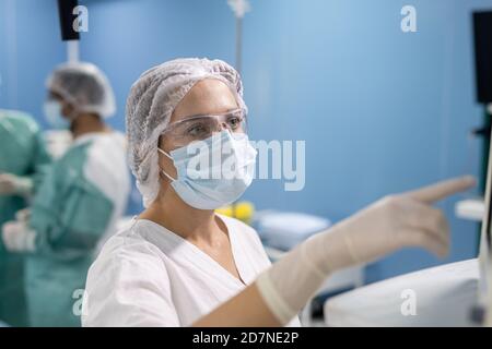 Giovane assistente del chirurgo in guanti, maschera e puntamento uniforme al display Foto Stock