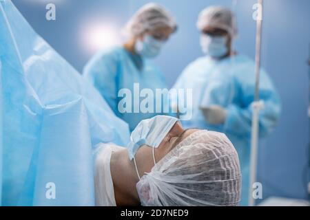 Paziente giovane in maschera e copricapo giacente sul tavolo operatorio contro i chirurghi Foto Stock