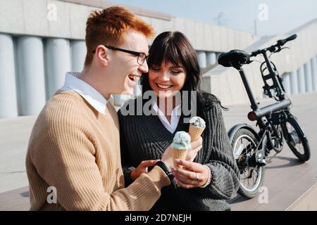 Uomo che si allaccia una donna che si siede all'aperto, tiene un gelato, parla, ride Foto Stock