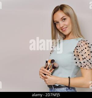 Concetto di trucco. Donna bionda sorridente che tiene spazzole cosmetiche, in posa su uno sfondo chiaro. Riprese in studio con spazio libero Foto Stock
