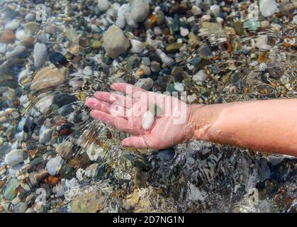 La mano delle donne tiene pietre marine nell'acqua di mare limpida. Acqua di mare cristallina attraverso la quale si possono vedere le pietre che giacciono sul fondo. Ecologia Foto Stock
