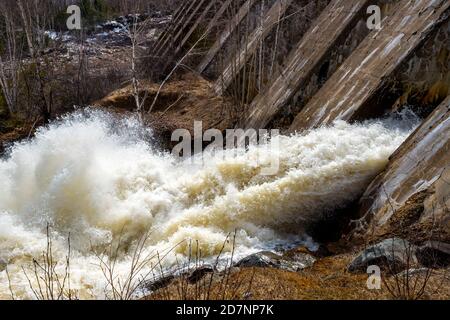 Acqua gushing dal fondo os una diga di calcestruzzo. L'acqua scorre rapidamente e schizza sul terreno. Alberi vuoti senza foglie presenti. Foto Stock