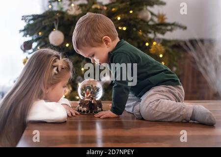 Sorella e fratello che guardano una palla di vetro con un Scena della nascita di Gesù Cristo in un bicchiere Palla su un albero di Natale Foto Stock