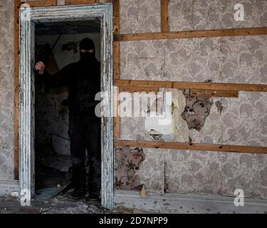 Un uomo che indossa una maschera/cappuccio in un edificio abbandonato. Sta tenendo un grande coltello, e sta in piedi nelle ombre in una porta. Foto Stock