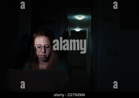 Freelancer femminile concentrato in occhiali seduti in camera oscura e. lavoro su progetto remoto su laptop in tarda serata a. casa Foto Stock