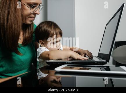 Vista laterale del bambino che gioca con la tastiera del computer portatile mentre si siede sulle ginocchia della madre che lavora a distanza in ufficio. Foto Stock
