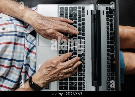 Vista dall'alto del ritaglio di un operatore remoto anonimo anziano che scrive sulla tastiera del laptop mentre si è seduti al tavolo sul posto di lavoro. Foto Stock