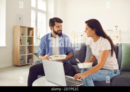 Una coppia allegra che mangia il cibo dalla consegna seduto davanti al computer portatile che parla e parla di film. Foto Stock